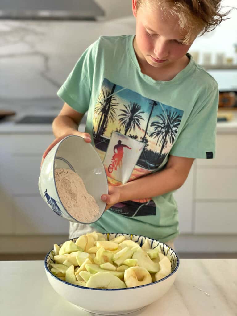 Boy adding flour and sugar mix to bowl of sliced apples