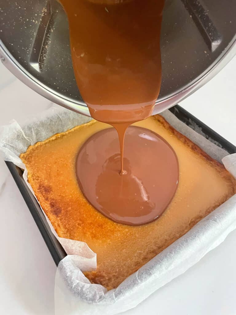 Pouring melted chocolate from Thermomix jug onto caramel slice. 