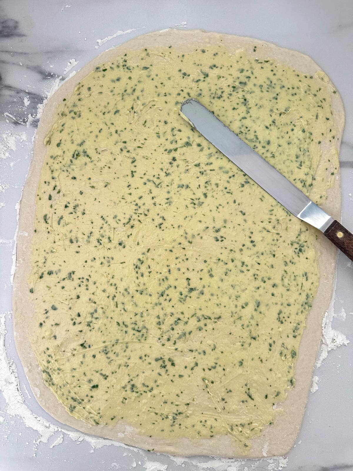 Knife spreading garlic butter over bread dough. 